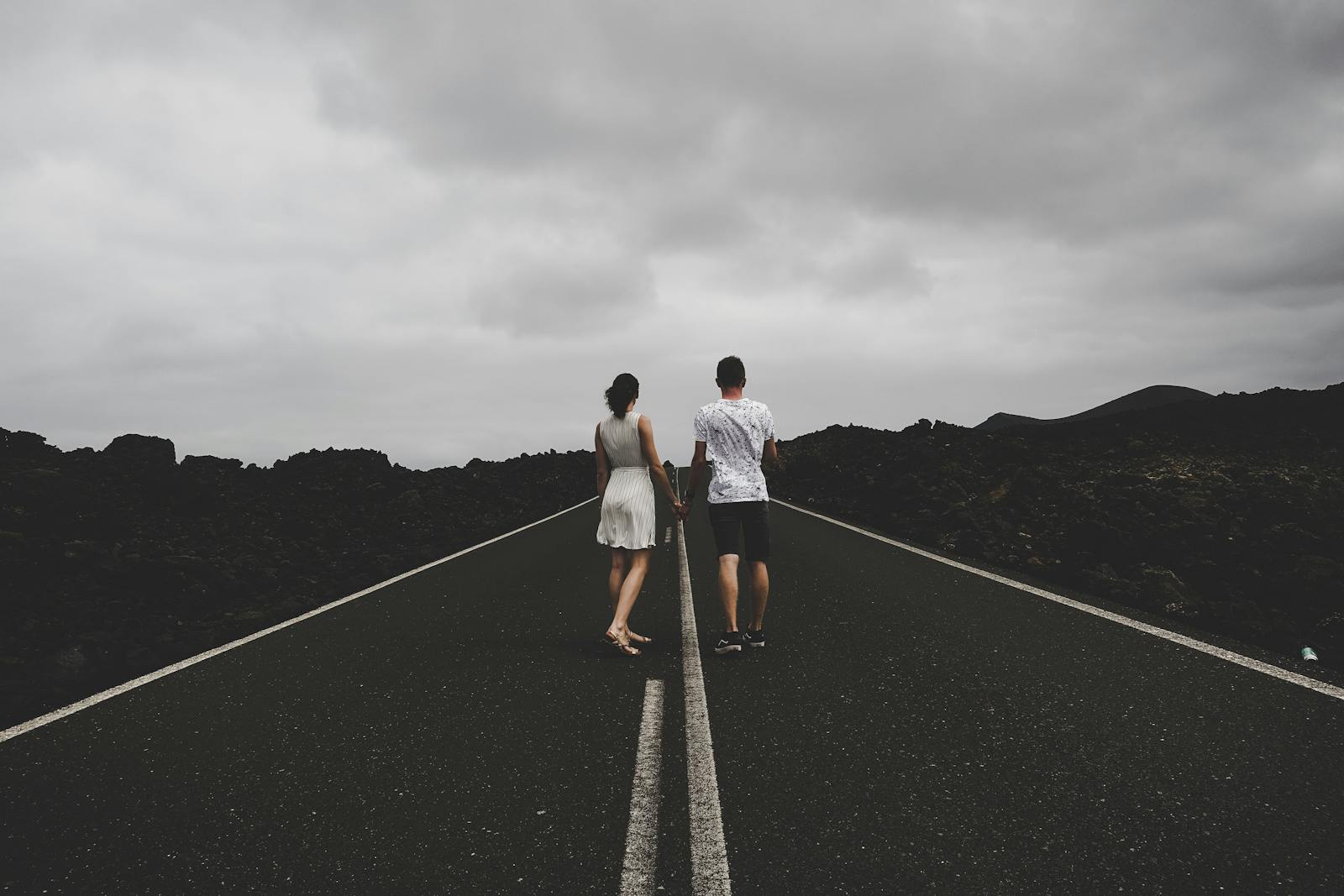 A couple walks hand in hand on an open road in Lanzarote, Spain, under a cloudy sky.