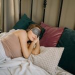 Woman resting peacefully in bed with a sleep mask, surrounded by cozy pillows in an inviting bedroom ambience.