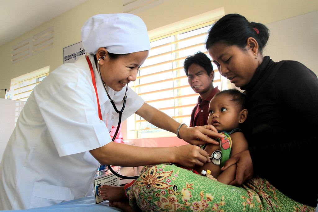 A nurse smiles while using a stethoscope to examine a baby with an ultra-rare genetic disorder sitting on their mother’s lap in a brightly lit clinic room. Another adult stands in the background, watching the interaction.