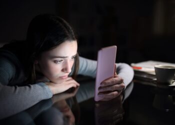 Sad woman checking phone at night at home, reflecting social media posting psychology and loneliness
