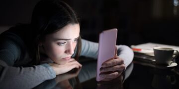 Sad woman checking phone at night at home, reflecting social media posting psychology and loneliness