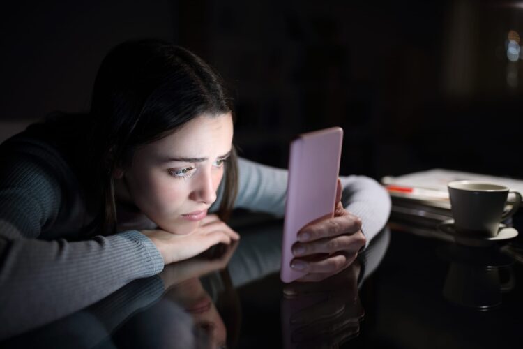 Sad woman checking phone at night at home, reflecting social media posting psychology and loneliness